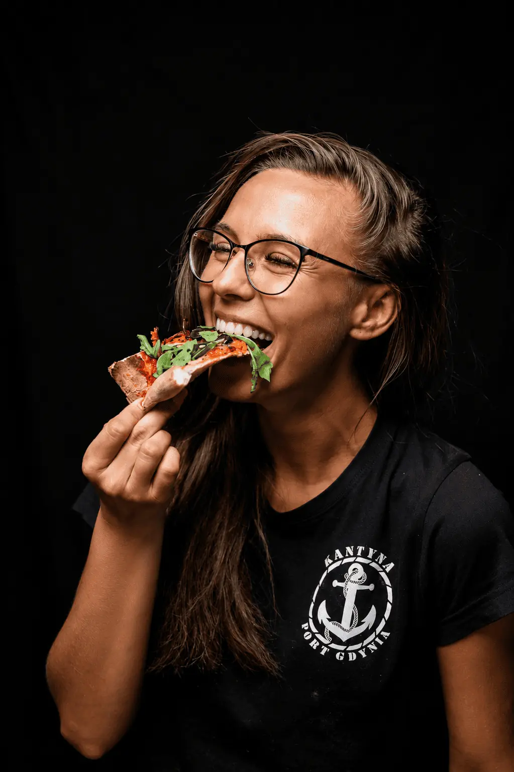 Woman with glasses smiling while taking a bite of a pizza slice against a black background, in a casual pose.