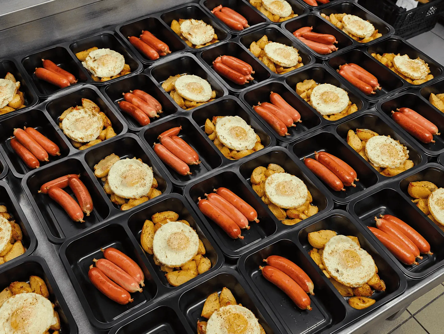Meal-prep trays arranged in rows, each with two sausages and a round breaded item on roasted potatoes in black containers.