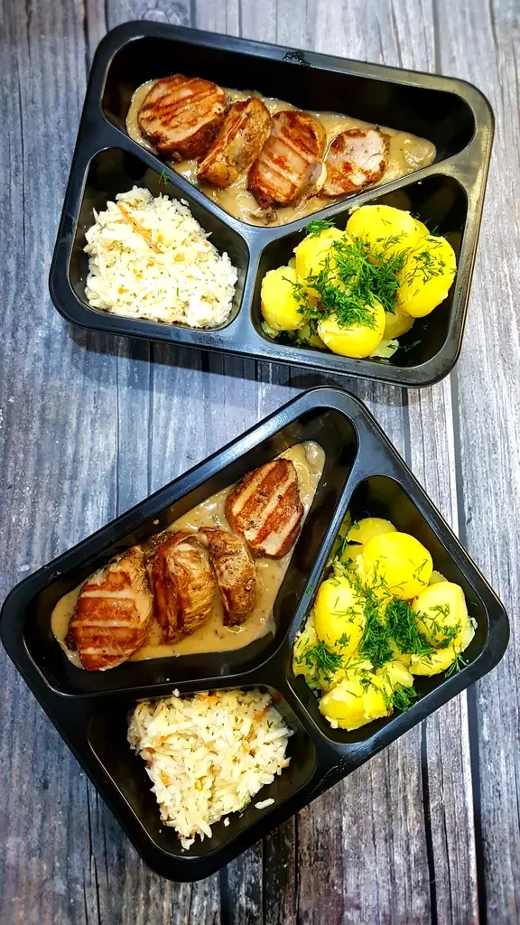 Two black, three-compartment meal trays on a wooden surface: pork medallions in creamy sauce, dill potatoes, and rice in each tray.