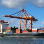 Container ship at a busy port with tall orange gantry cranes and stacked colorful shipping containers beside the water