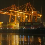 Night scene of a container ship named northern defender docked at a port with tall orange gantry cranes and stacked cargo containers reflecting on calm water