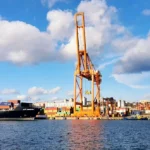 Port of loading with bright orange cranes stacked shipping containers and a large cargo ship by calm water under a blue sky
