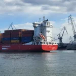 Red container ship at a port with stacked colorful cargo containers and large cranes nearby in a busy harbor setting